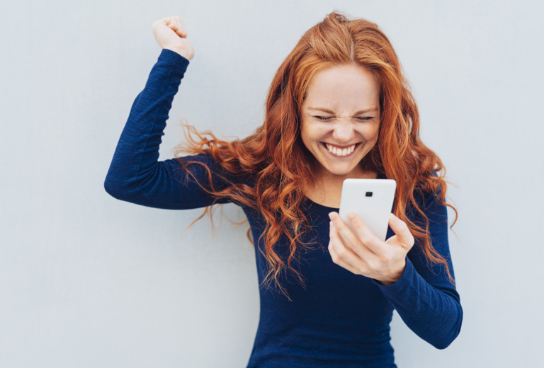 junge Frau schaut jubelnd auf ihr Smartphone und hebt eine Hand in die Luft